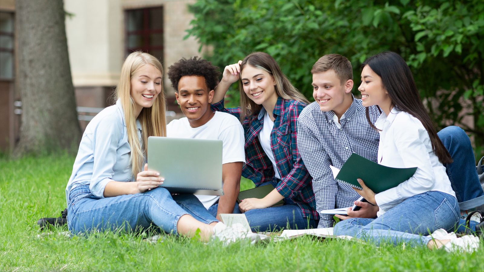 Das Bild zeigt mehrere diverse junge Menschen, die mit Lernmaterialien auf einer Wiese sitzen und zu einem Thema diskutieren.