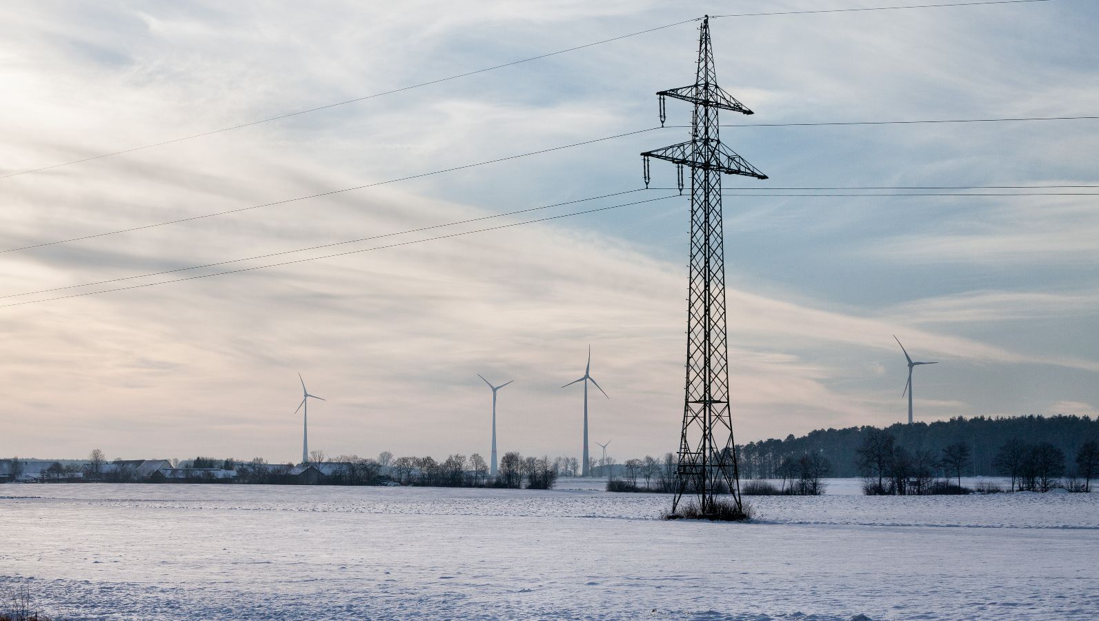 Das Bild zeigt einen Starkstrommast und im Hintergrund Windräder auf einem verschneiten Feld bei einer Winterdämmerung.