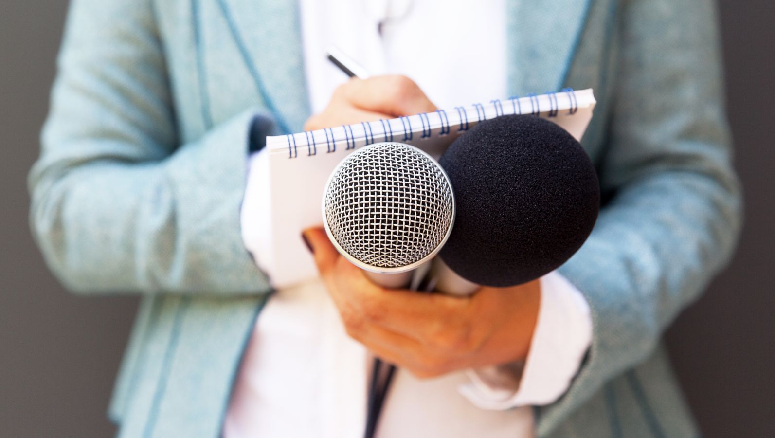 Zu sehen ist in Nahaufnahme das Arbeitsmaterial von Medienschaffenden - Notizblock, Stift, Mikrofone - in den Händen einer weiblichen Person.